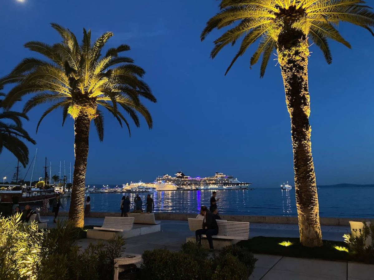 Kuvassa satama-alue, jossa iltahämärässä valaistut palmut. Merellä näkyy risteilyalus valoineen.