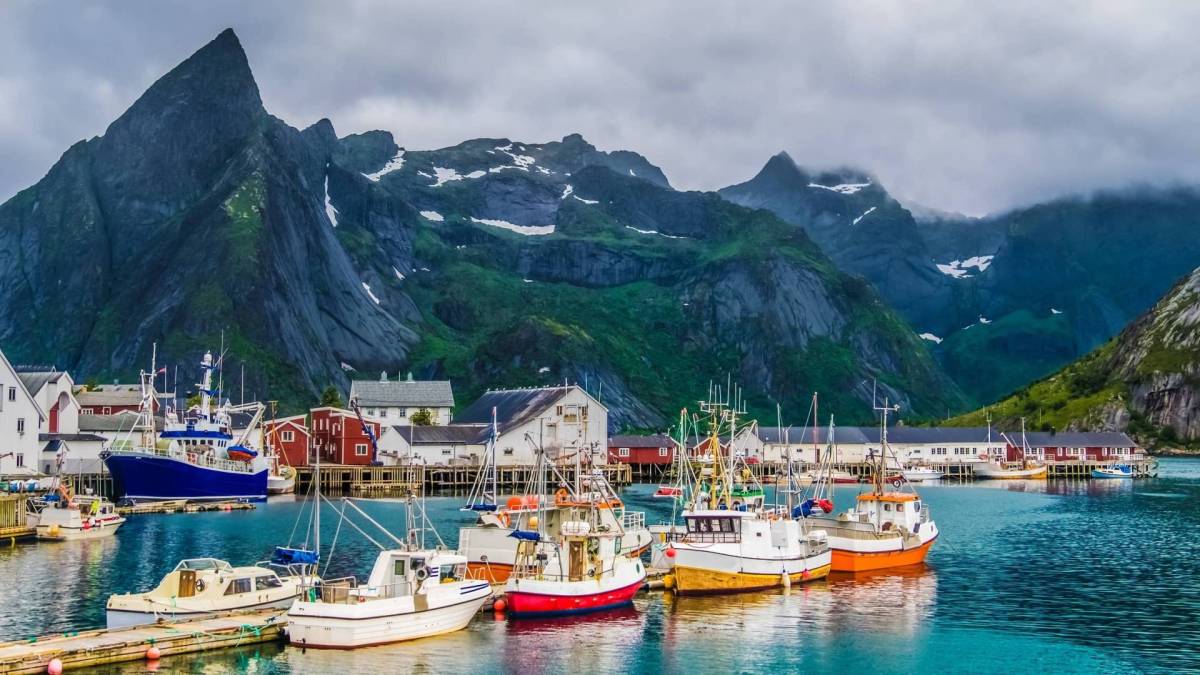 Kuvassa norjalaiskylä, jossa pieni satama täynnä veneitä ja taustalla kohoaa vuoristot. 