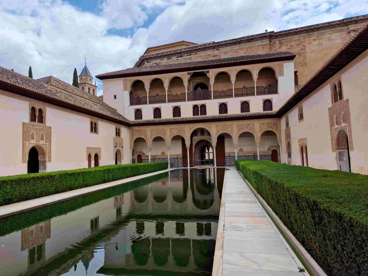Alhambran palatsi. Vaalea rakennus, jonka edessä laatikonmallisesti leikatut pensaat ja pieni vesialue. 