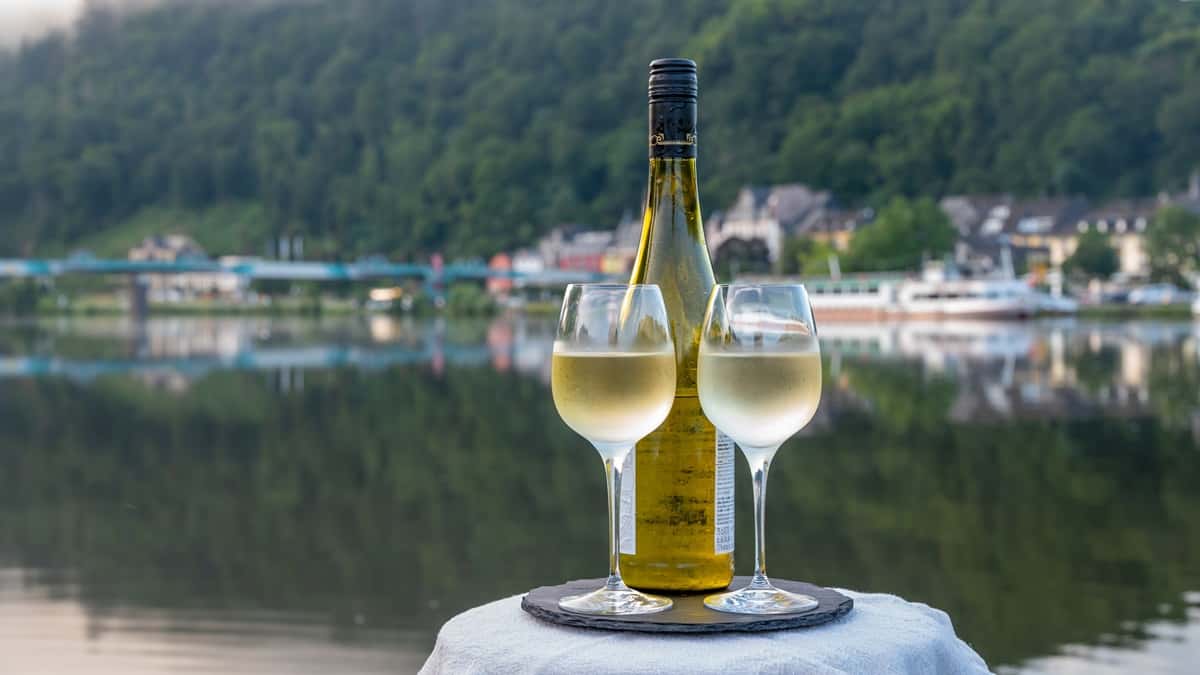 Jokiristeilyt Euroopassa ovat kulinaristia elämyksiä. Mosel on tunnettu hyvistä rieslingeistä Jokiristeilyt Euroopassa ovat täynnä ruoka- ja juomakulttuuria. Kuvassa kaksi viinilasia ja valkoviinipullo, taustalla Moseljoen vihreää maisemaa.