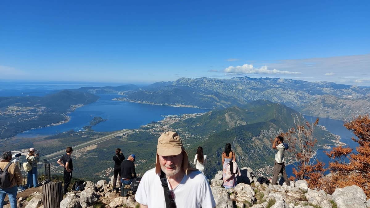 Mies hattu päässään vuoren rinteellä. Taustalla merimaisema ja vuoristoa.